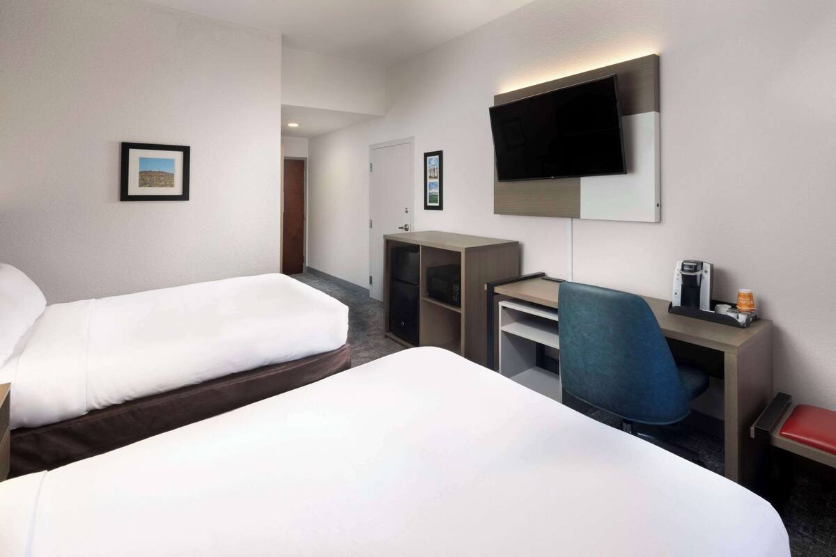 A hotel room with two beds, a desk, a chair, a TV, and a small table with a coffee maker.