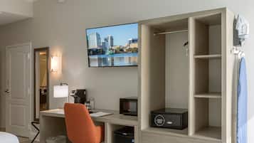 Habitación de hotel con cama, escritorio con computadora, silla, televisor mostrando un paisaje urbano, caja fuerte y armario.
