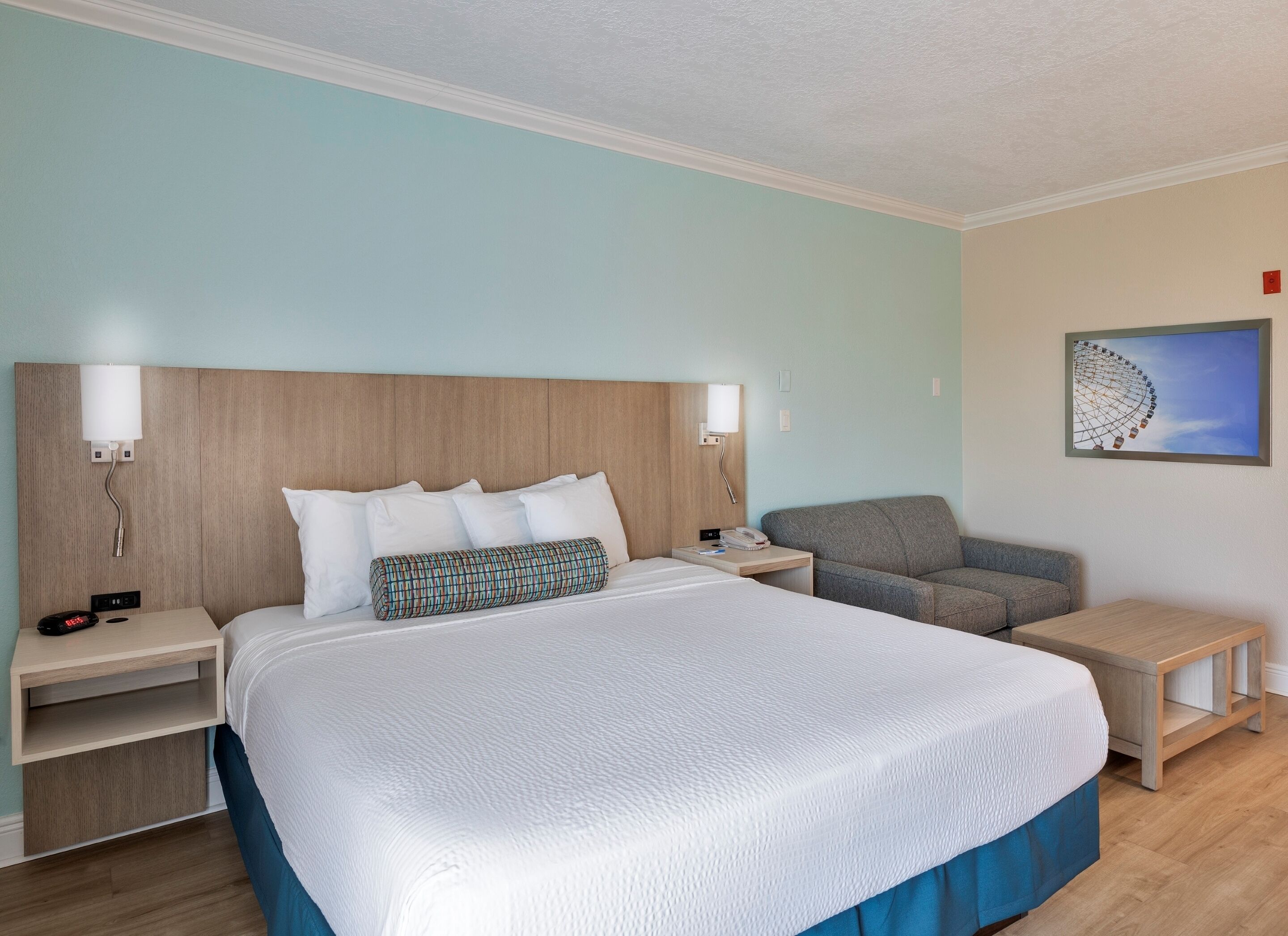 A hotel room with a large bed, a grey armchair, a small table, and a wall-mounted artwork.