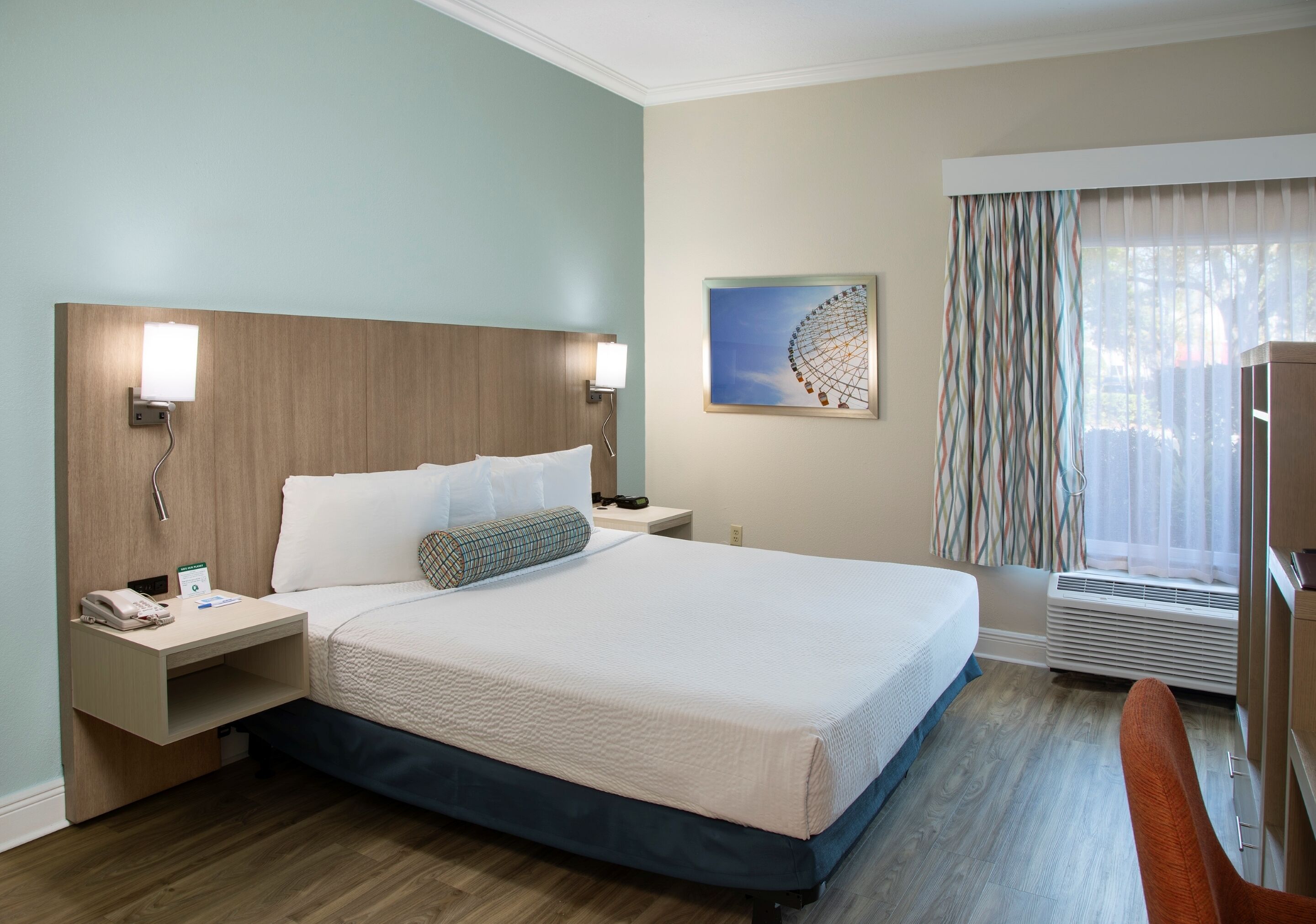 Une chambre d’hôtel avec un grand lit, des tables de chevet, une lampe fixée au mur, une fenêtre avec des rideaux et une œuvre d’art décorative au mur.
