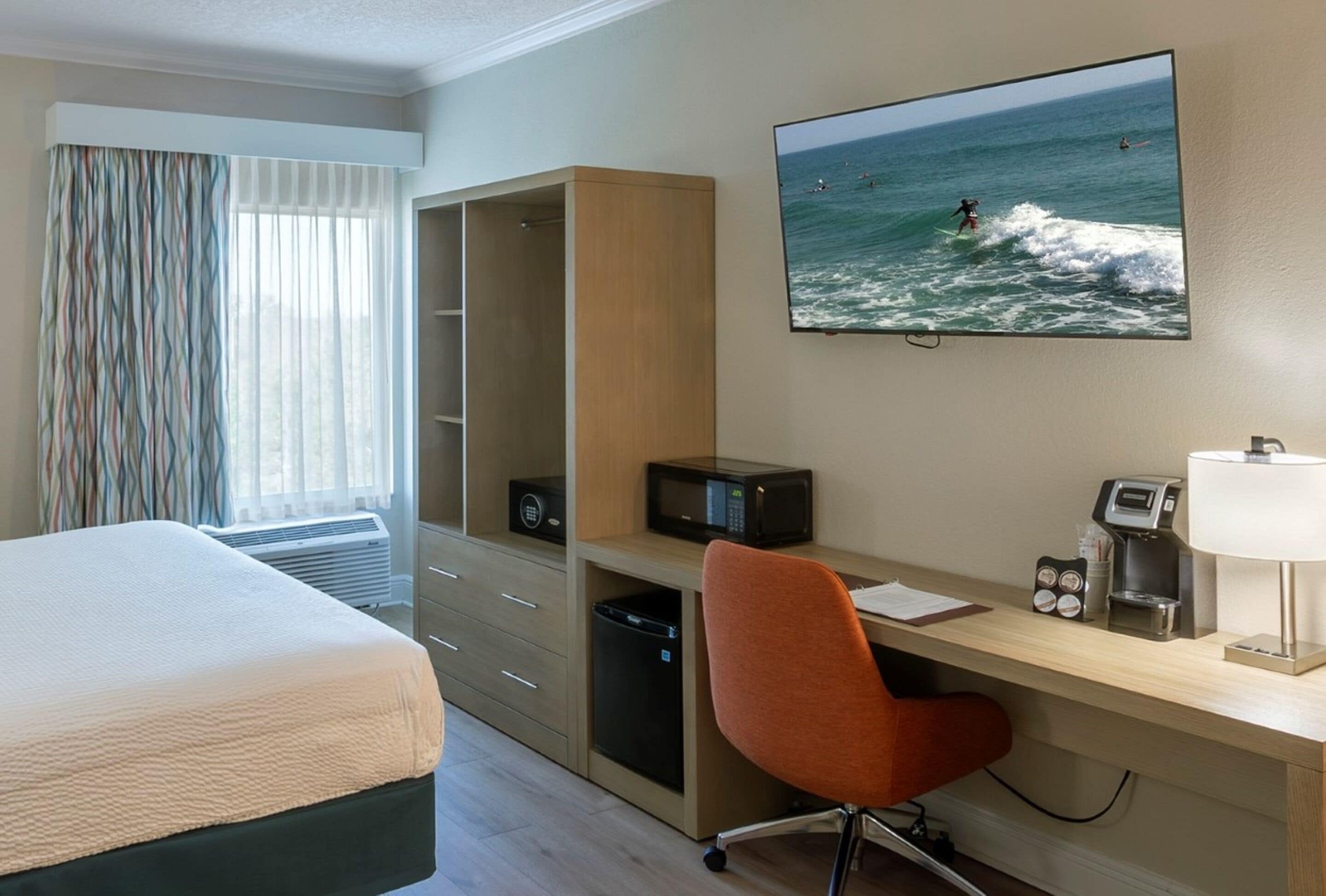 Une chambre d’hôtel avec un lit, un bureau et une chaise, un téléviseur diffusant une émission sur le surf, et une fenêtre avec des rideaux.