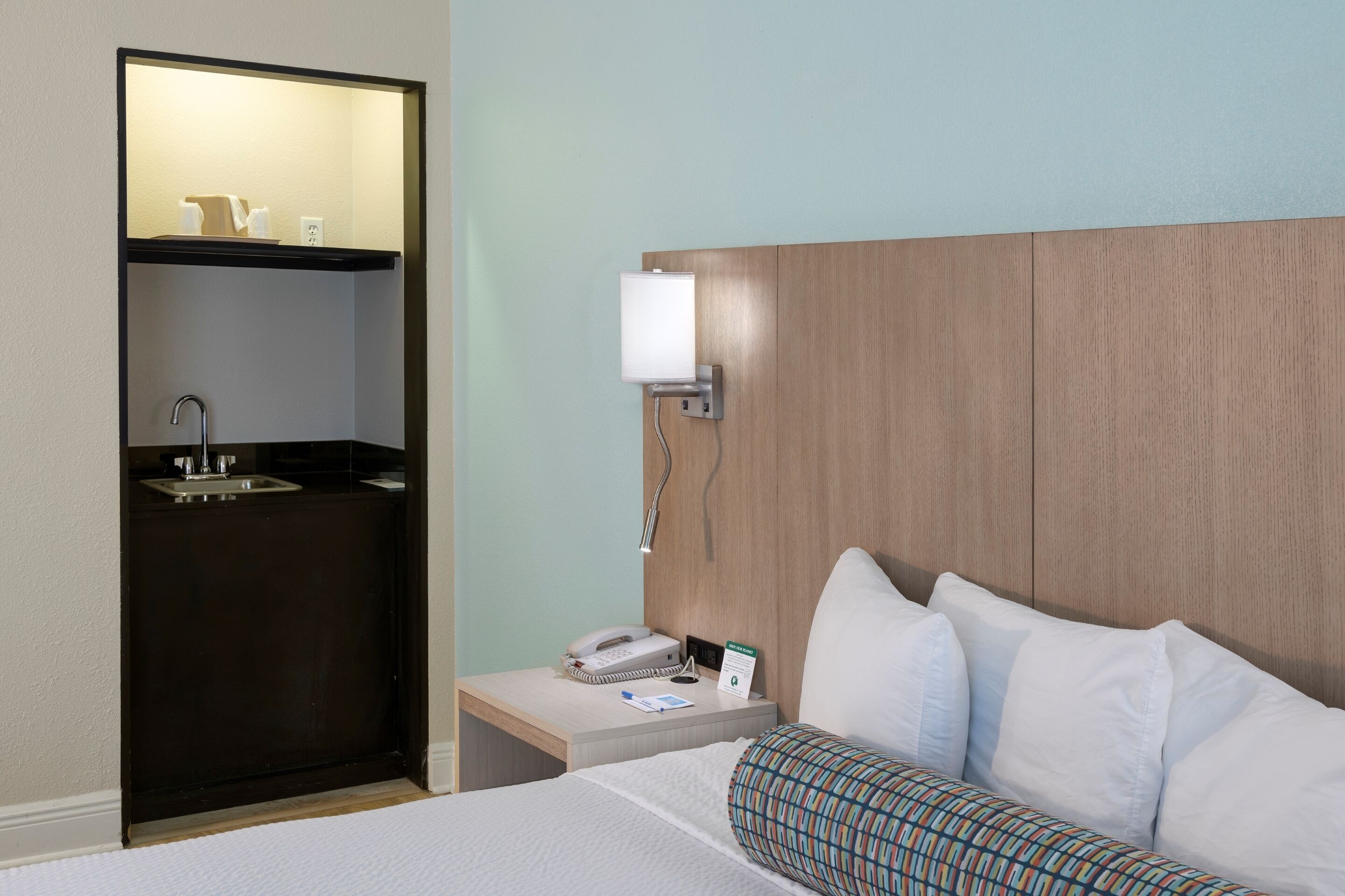 Quarto de hotel com cama, mesa de cabeceira com telefone e luminária, banheiro pequeno com pia e espelho, e cabeceira de madeira.