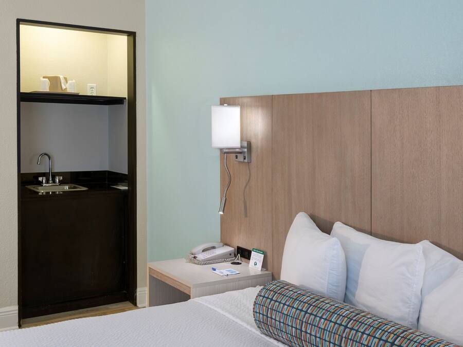 Hotelkamer met een bed, nachtkastje met telefoon en lamp, kleine badkamer met wastafel en spiegel, en een houten hoofdbord.