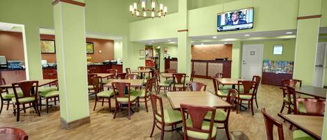 A restaurant with multiple wooden tables and chairs, a kitchen area, and a television mounted on the wall.