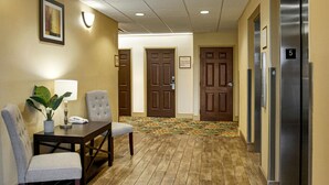 A hotel lobby with a reception desk, two armchairs, a potted plant, a telephone, and a carpeted floor. There are two doors in the background, one with a sign indicating room numbers.