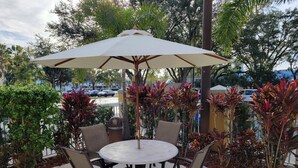 An outdoor patio with a round table and chairs, surrounded by lush greenery and a large umbrella.