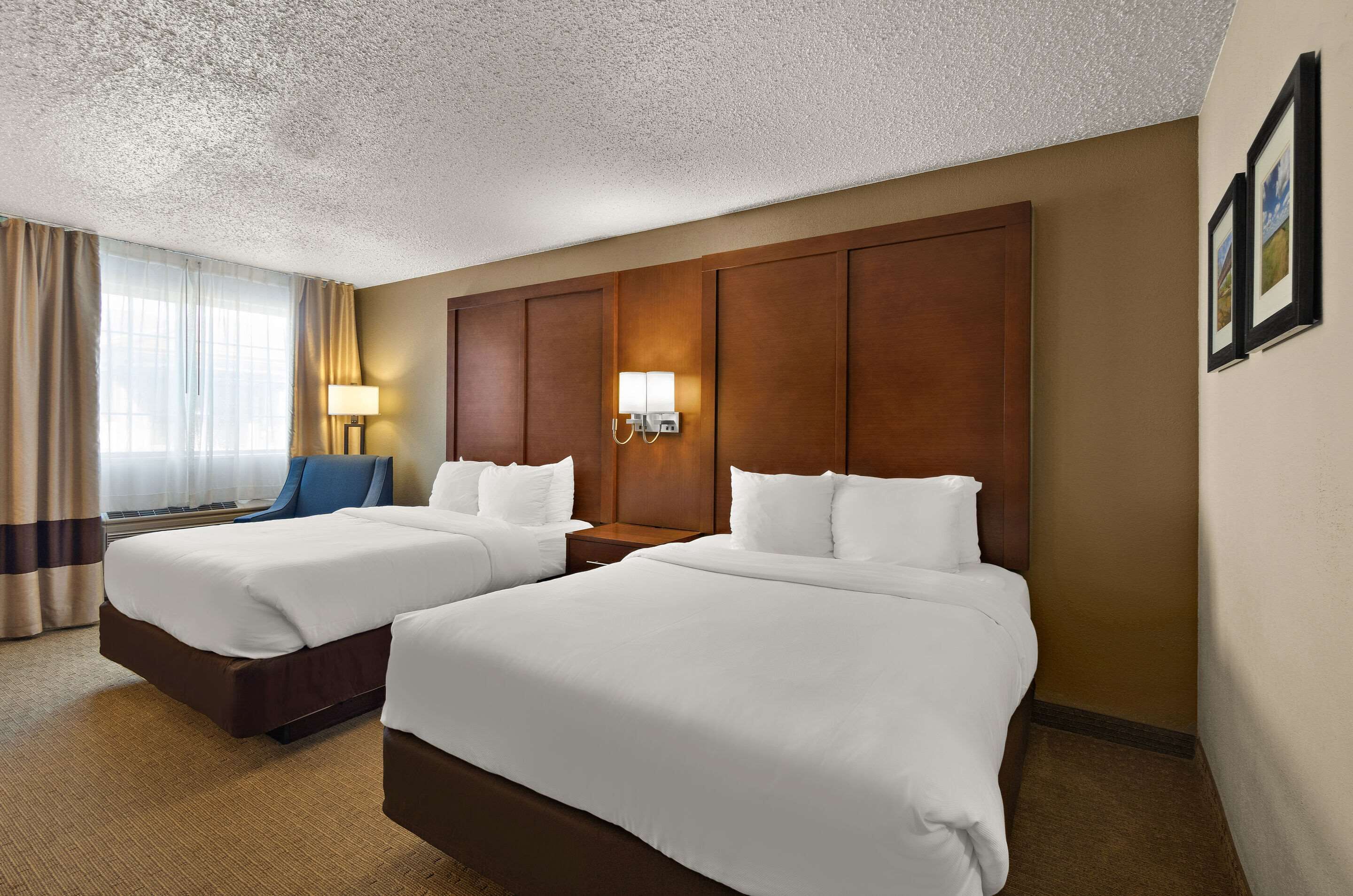 A hotel room with two beds, a wooden headboard, a nightstand, a lamp, and a window with curtains.