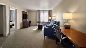 Habitación de hotel con cama, escritorio, silla, televisor, lámpara y vista a la ciudad a través del ventanal.
