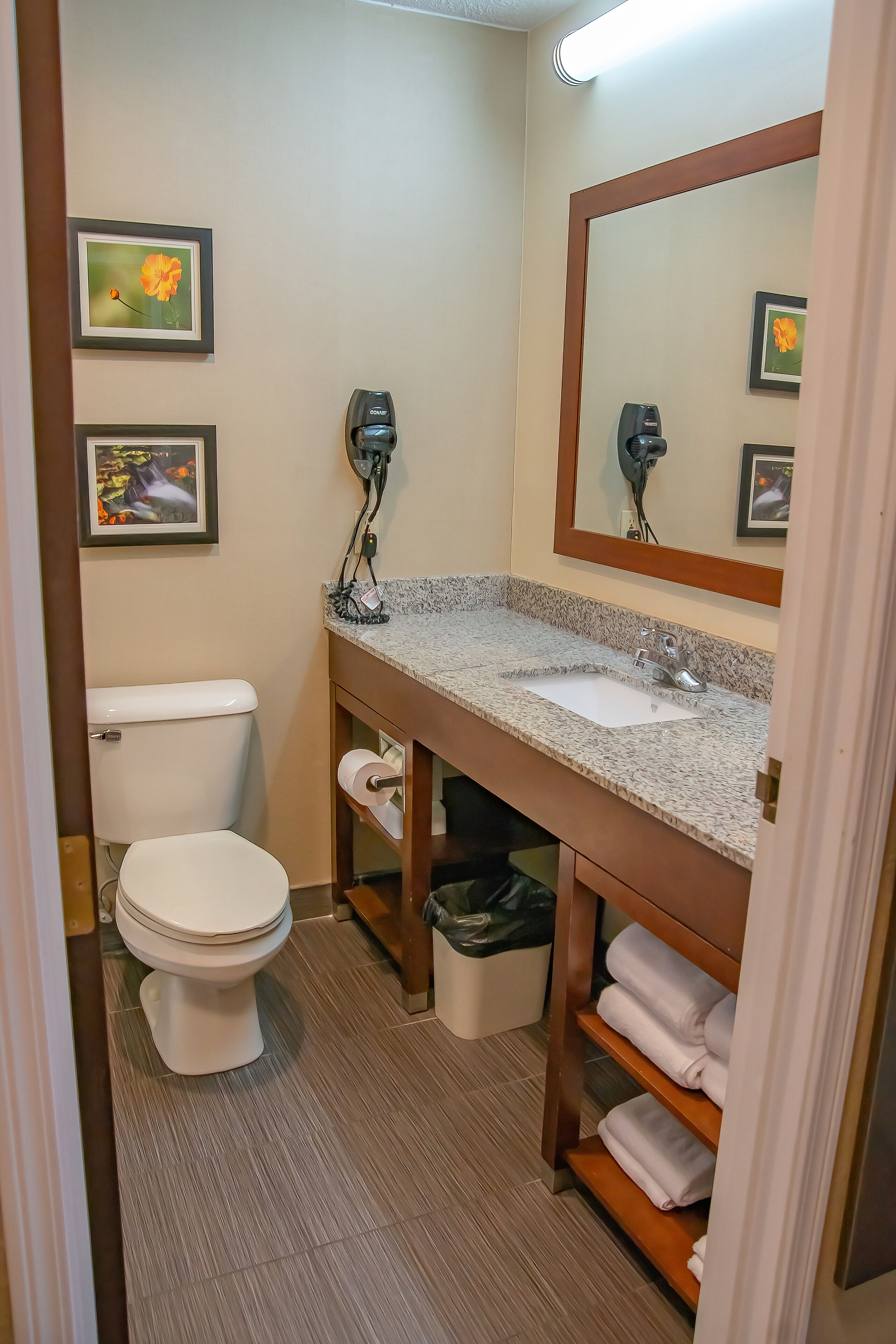 A compact bathroom with a toilet, sink, and mirror. There are framed pictures on the wall, a hairdryer, and a trash bin.