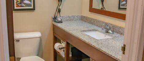 A compact bathroom with a toilet, sink, and mirror. There are framed pictures on the wall, a hairdryer, and a trash bin.