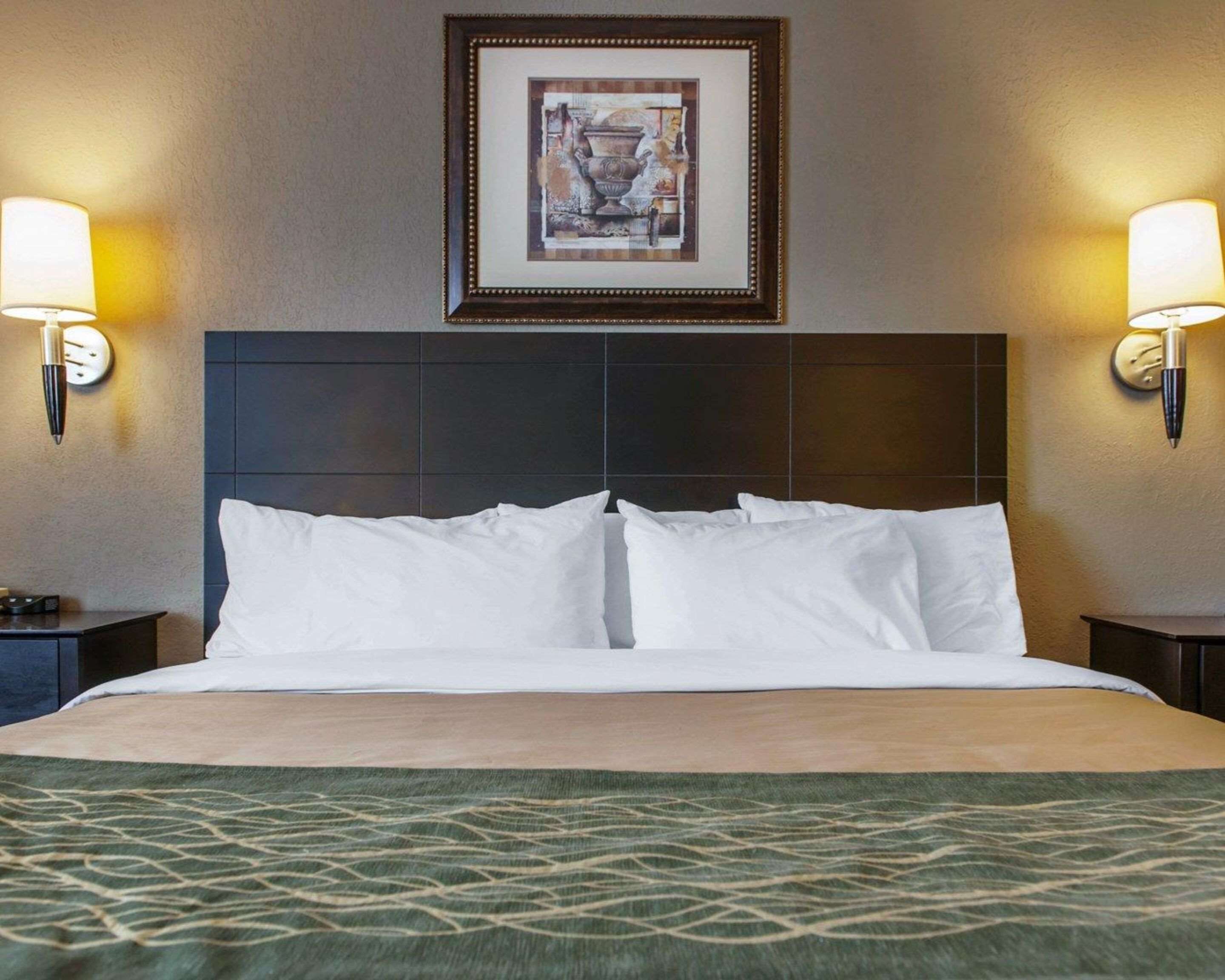 Una camera d'albergo con un letto grande, due lampade ai comodini, un quadro appeso alla parete e un comodino con una penna e una sveglia.