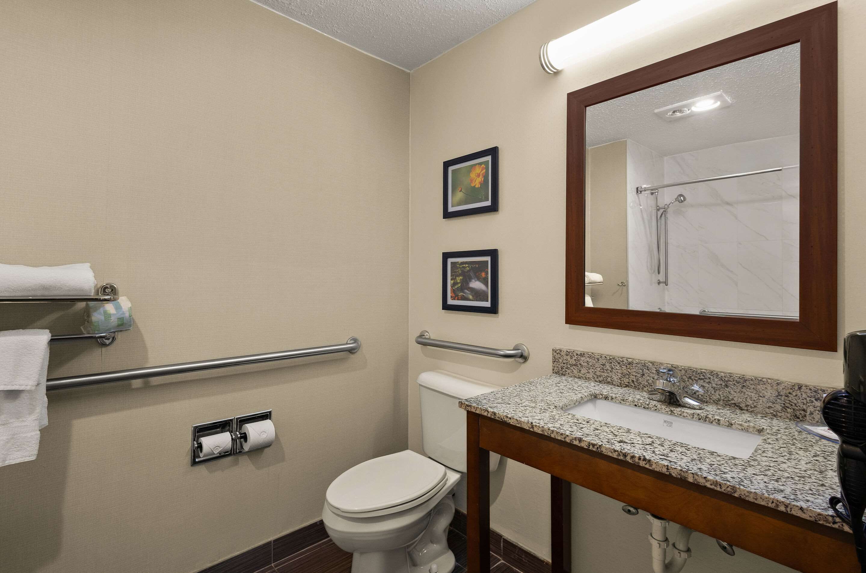 A small bathroom with a toilet, sink, and mirror. There are two framed pictures on the wall, a towel rack, and a handrail.