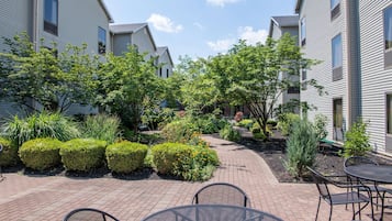 A courtyard with a brick pathway, metal tables and chairs, and various shrubs and plants.