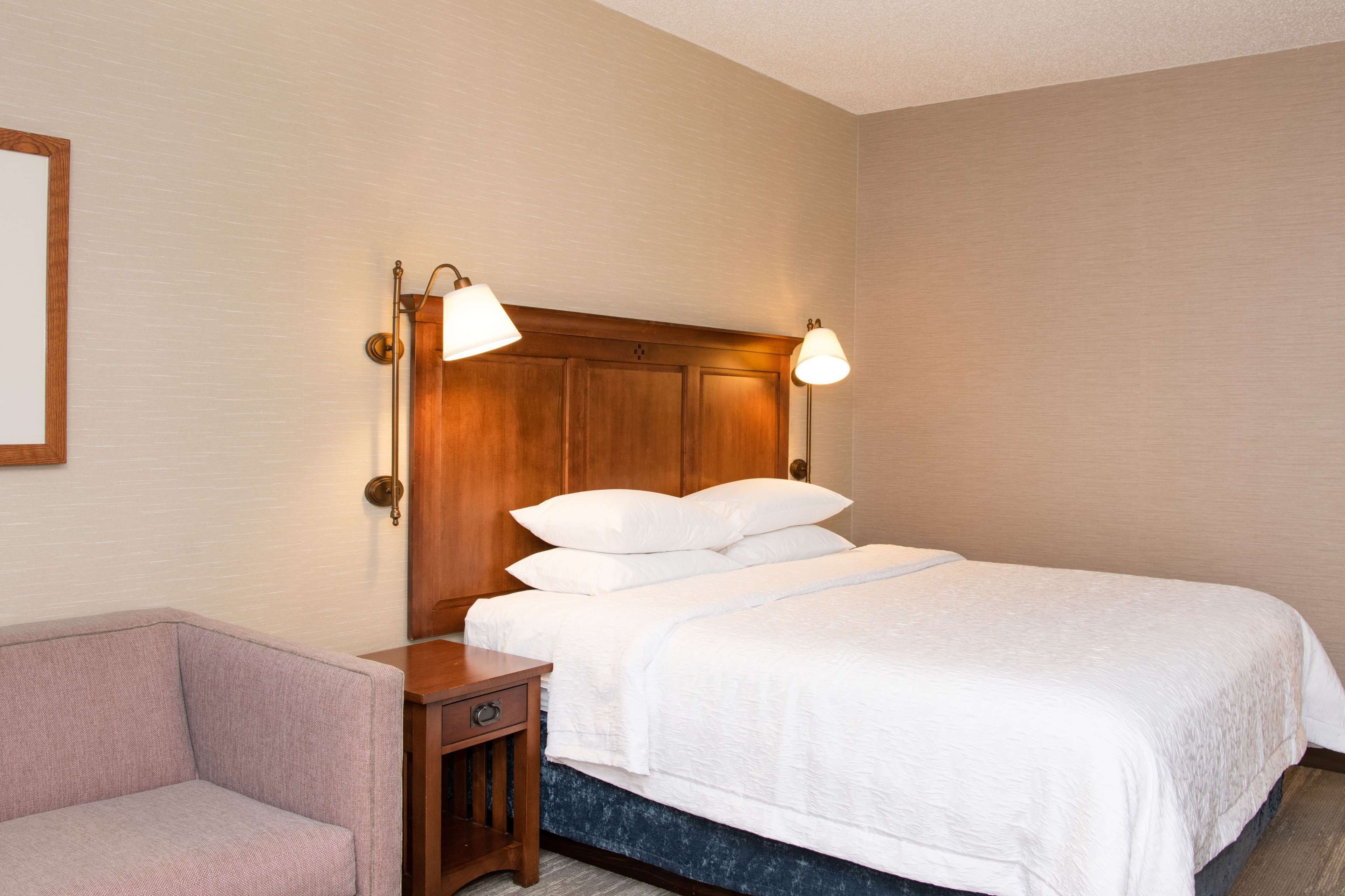 Una habitación de hotel con cabecera de madera, una cama con ropa blanca, dos lámparas de noche, una mesita de noche y una pequeña silla.