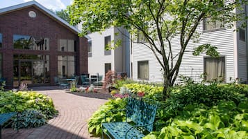 A courtyard with a brick building, a white building, and a tree. There are green plants, a blue bench, and a table with chairs.
