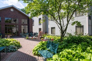 A courtyard with a brick building, a white building, and a tree. There are green plants, a blue bench, and a table with chairs. - Hampton Inn & Suites Rochester/Victor (Victor)