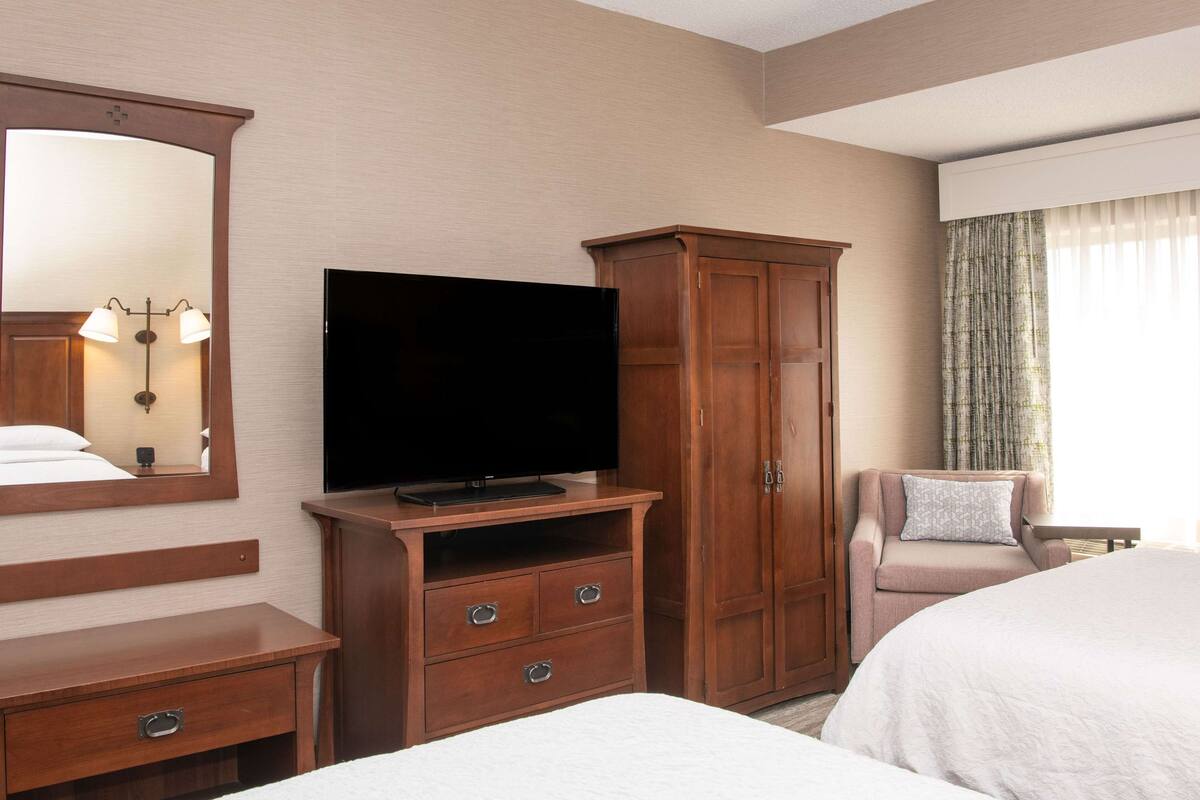 A hotel room with a bed, a TV on a wooden stand, a wardrobe, a chair, and a large mirror.