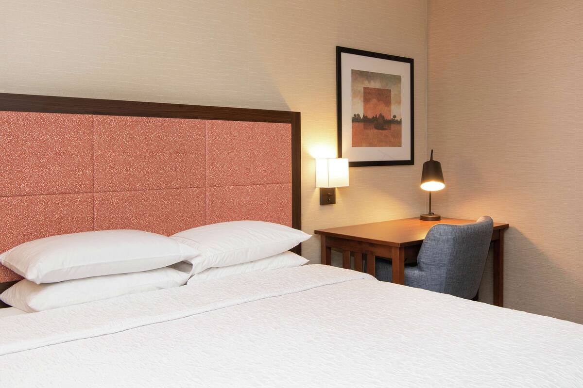 A hotel room with a large bed, a desk with a chair, a wall-mounted lamp, and a framed picture on the wall.