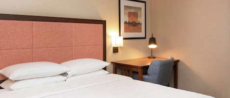A hotel room with a large bed, a desk with a chair, a wall-mounted lamp, and a framed picture on the wall.