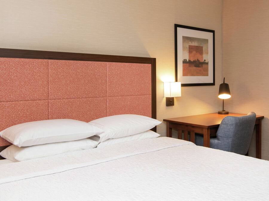 A hotel room with a large bed, a desk with a chair, a wall-mounted lamp, and a framed picture on the wall.