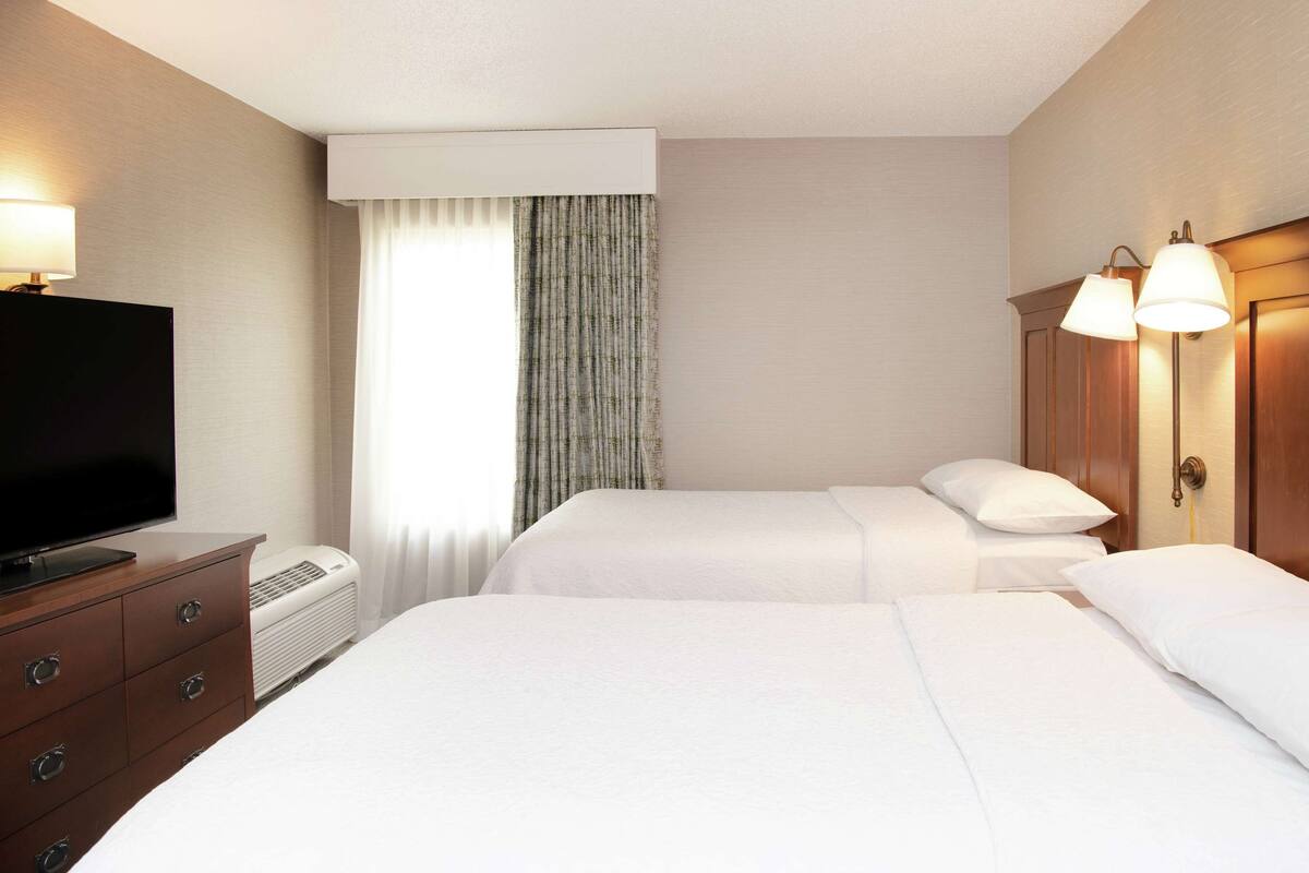 A hotel room with two beds, a TV, a dresser, and a window with curtains.