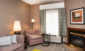 A hotel room with a sofa, armchair, table, lamp, window with curtains, fireplace, and a framed picture on the wall.