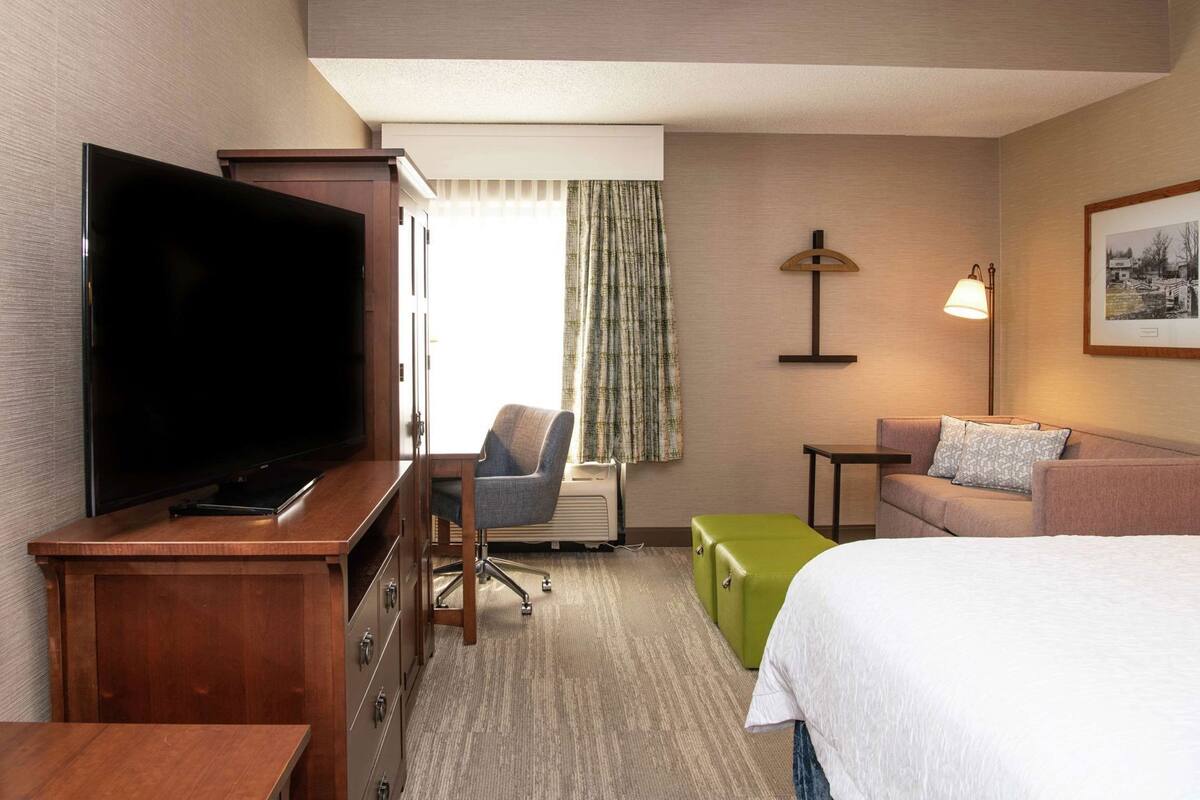 A hotel room with a bed, a television, a desk, a chair, a sofa, a lamp, and a picture on the wall.