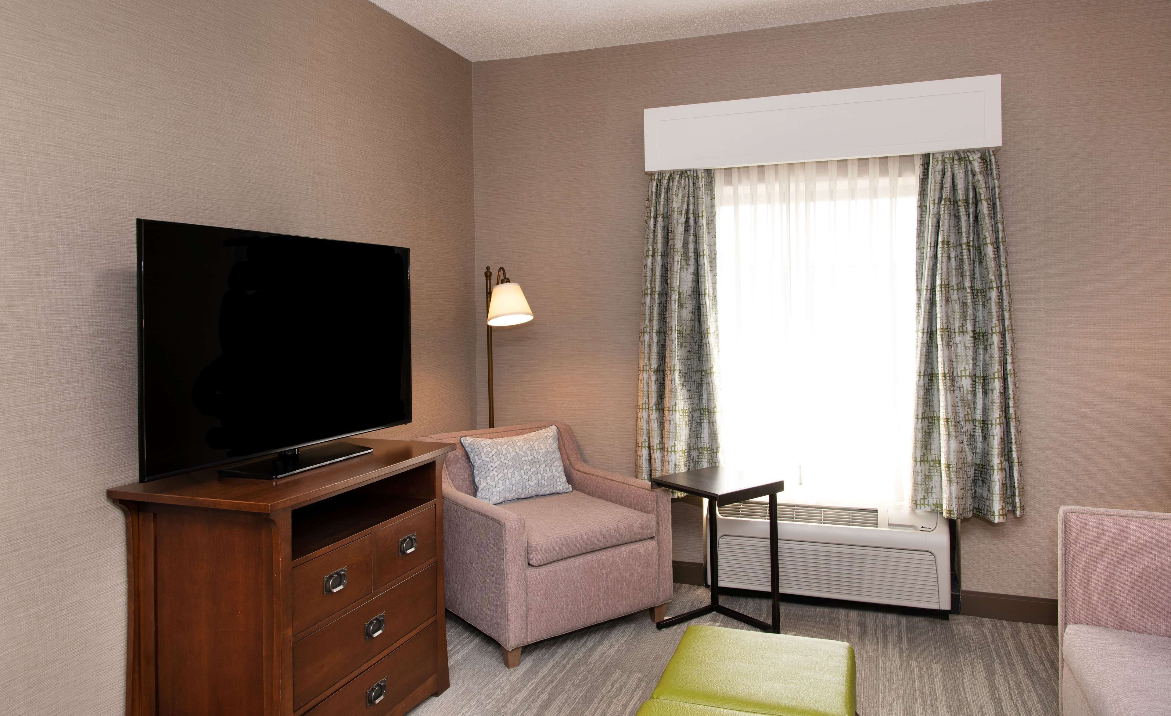 Ein Hotelzimmer mit Flachbildfernseher, einer Holzkommode mit Schubladen, einem rosa Sessel, einem kleinen Tisch und einem Fenster mit gemusterten Vorhängen.