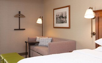 Habitación de hotel con cama, una silla verde, sofá, una mesita y dos lámparas de pared.