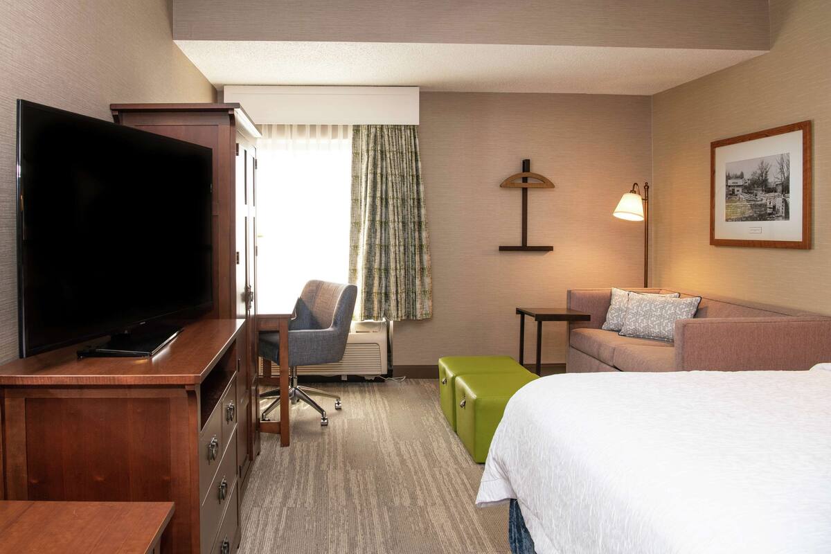 A hotel room with a flat-screen TV, a desk, a chair, a sofa, a bed, a lamp, and a framed picture on the wall.