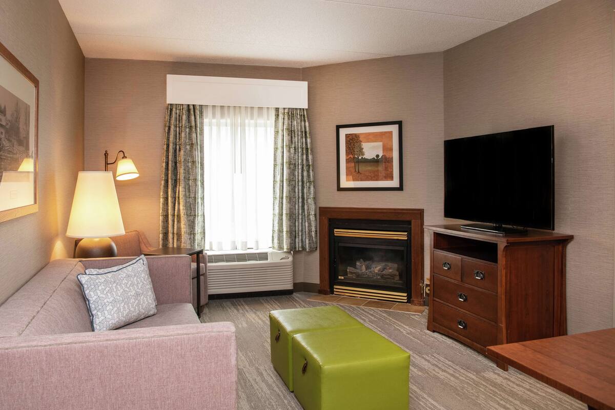 A hotel room with a sofa, a green ottoman, a television, a fireplace, and a window with curtains.