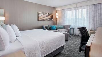 Habitación de hotel con una cama grande, un sofá, un escritorio y una silla. Se ve una ventana con cortinas, una lámpara y un cuadro en la pared.