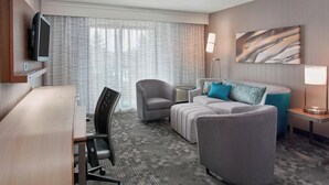 Hotelkamer met een wandgemonteerde flatscreen-tv, een grijze bank met blauwe kussens, een grijze fauteuil, een klein bijzettafeltje met een lamp en een groot raam met dunne gordijnen.