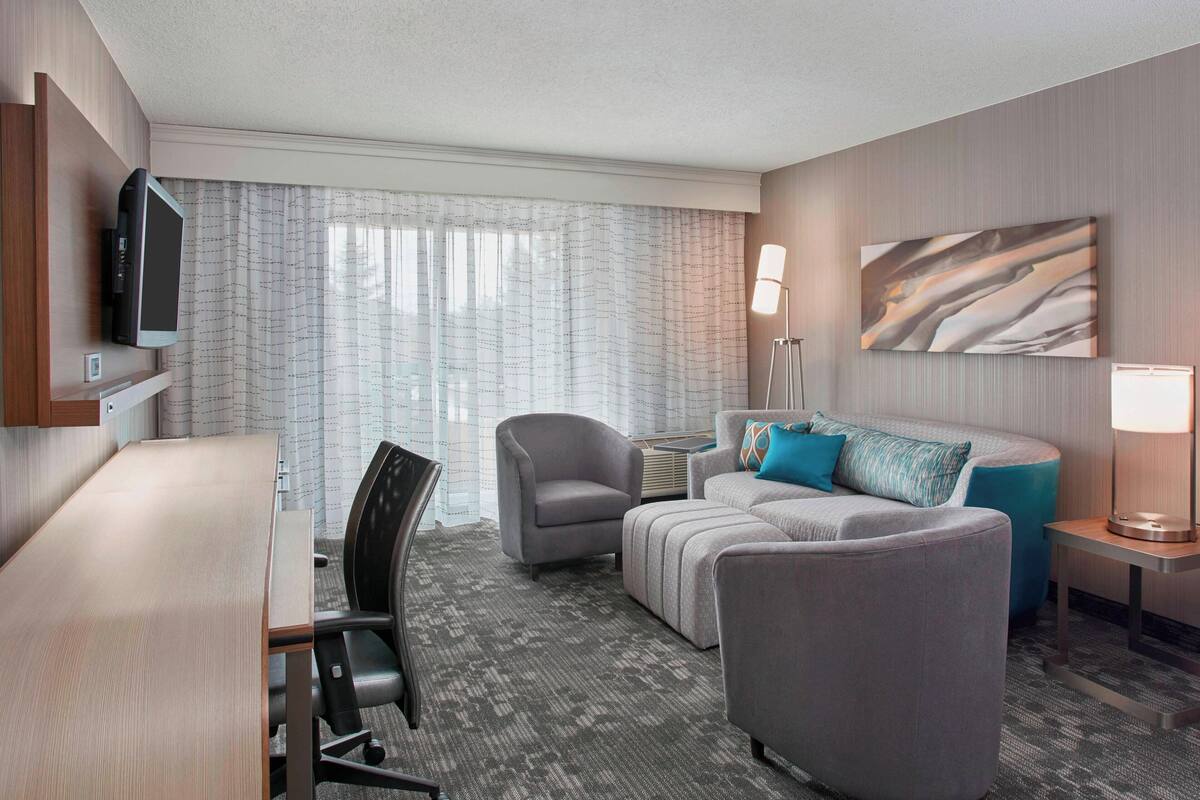 Hotelkamer met een wandgemonteerde flatscreen-tv, een grijze bank met blauwe kussens, een grijze fauteuil, een klein bijzettafeltje met een lamp en een groot raam met dunne gordijnen.