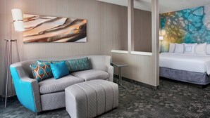 Een hotelkamer met een grijze bank, een klein bijzettafeltje, een bed met wit beddengoed en een groot abstract schilderij aan de muur.