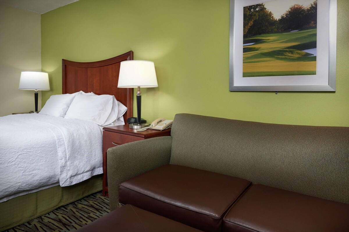 Una habitación de hotel con una cama, dos lámparas de noche, un teléfono, un cuadro de paisaje enmarcado y un sofá de cuero marrón.