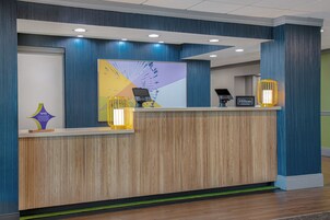 Un mostrador de recepción de hotel con diseño moderno, que incluye un panel frontal de madera, una gran obra de arte abstracta y un quiosco digital.