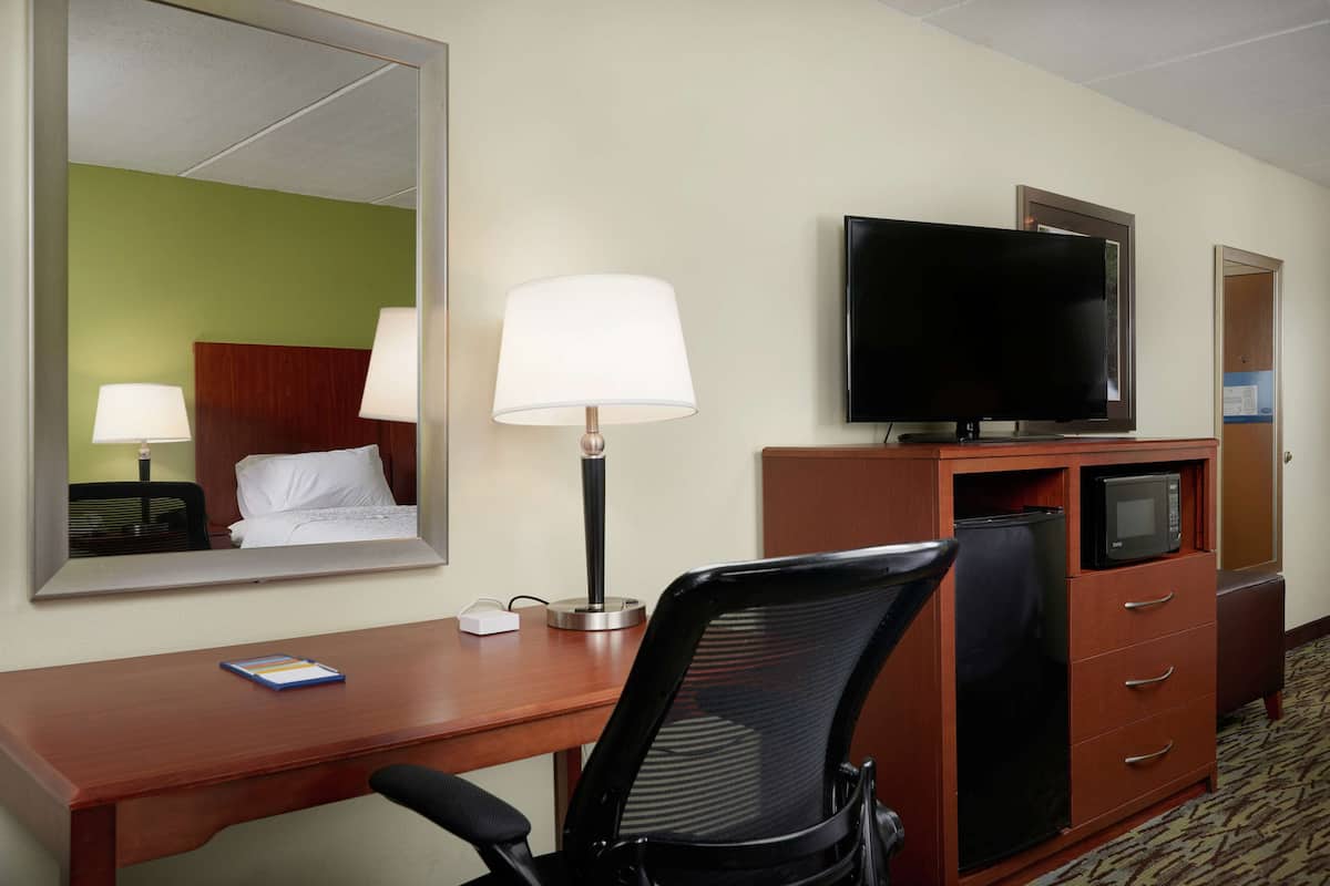 Ett hotellrum med ett skrivbord i trä, en svart kontorsstol, en platt-TV, en mikrovågsugn, en spegel och en säng med vita sängkläder.