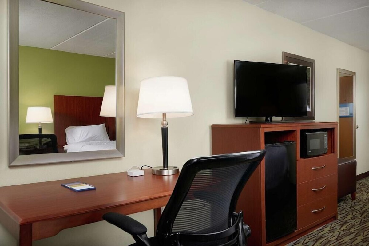 Habitación de hotel con escritorio, silla, televisor, lámpara y un espejo que refleja la habitación.