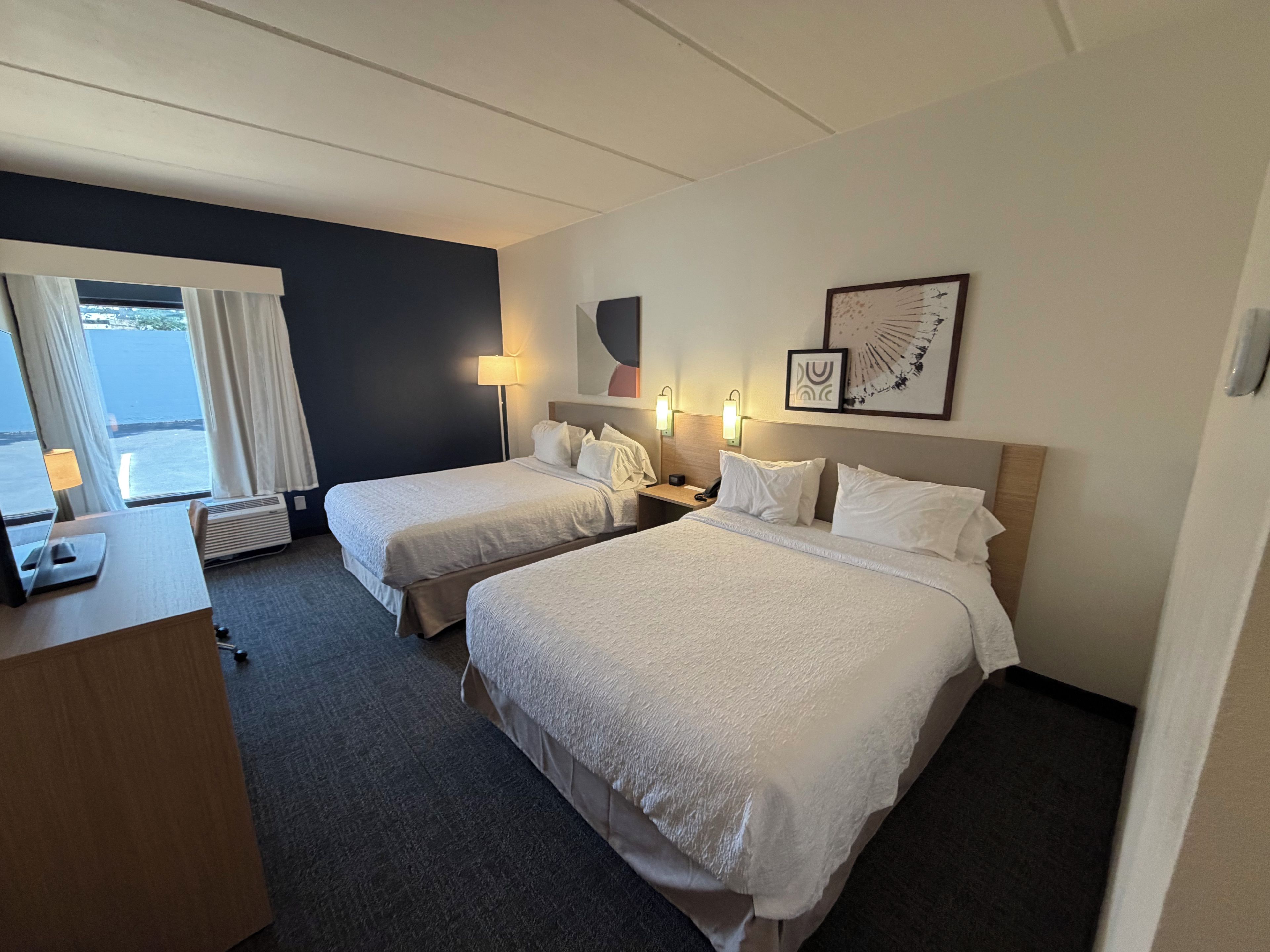 Habitación de hotel con dos camas, un escritorio, una lámpara y vista al mar.