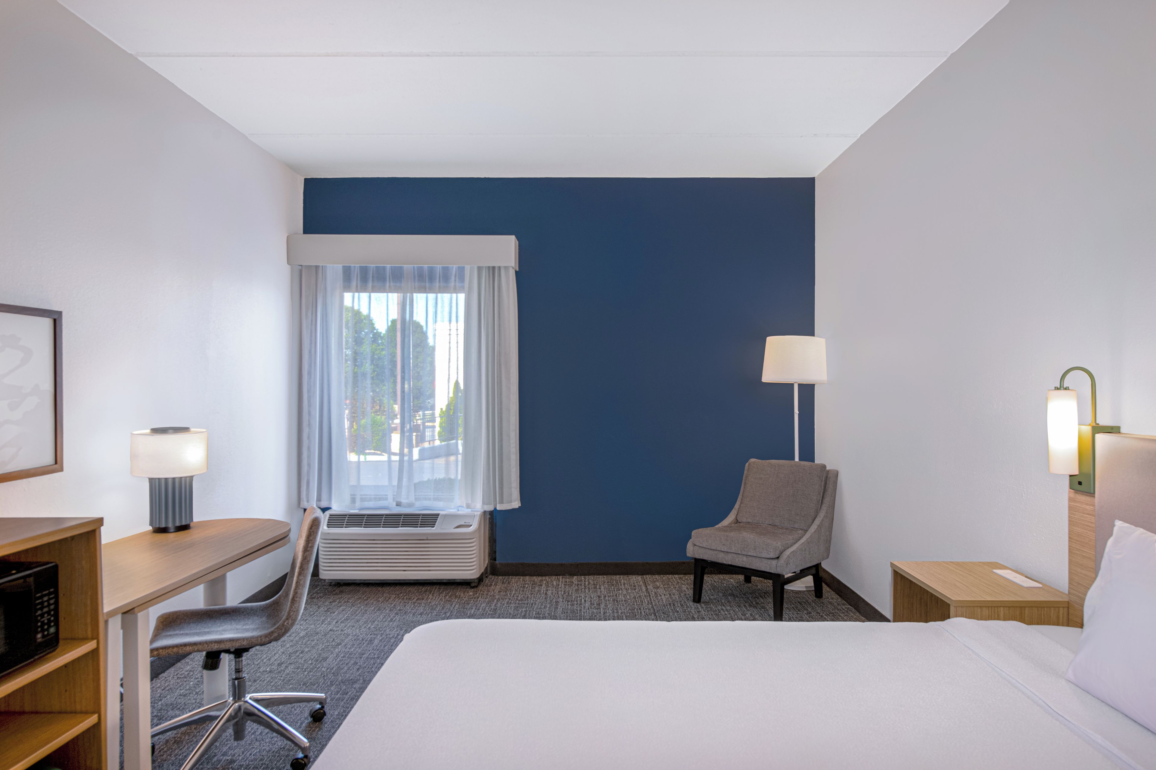 A hotel room with a bed, a desk, a chair, a window with curtains, and a wall-mounted lamp.