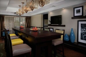 A modern hotel dining area with a central bar, cushioned seating, and a large TV on the wall.