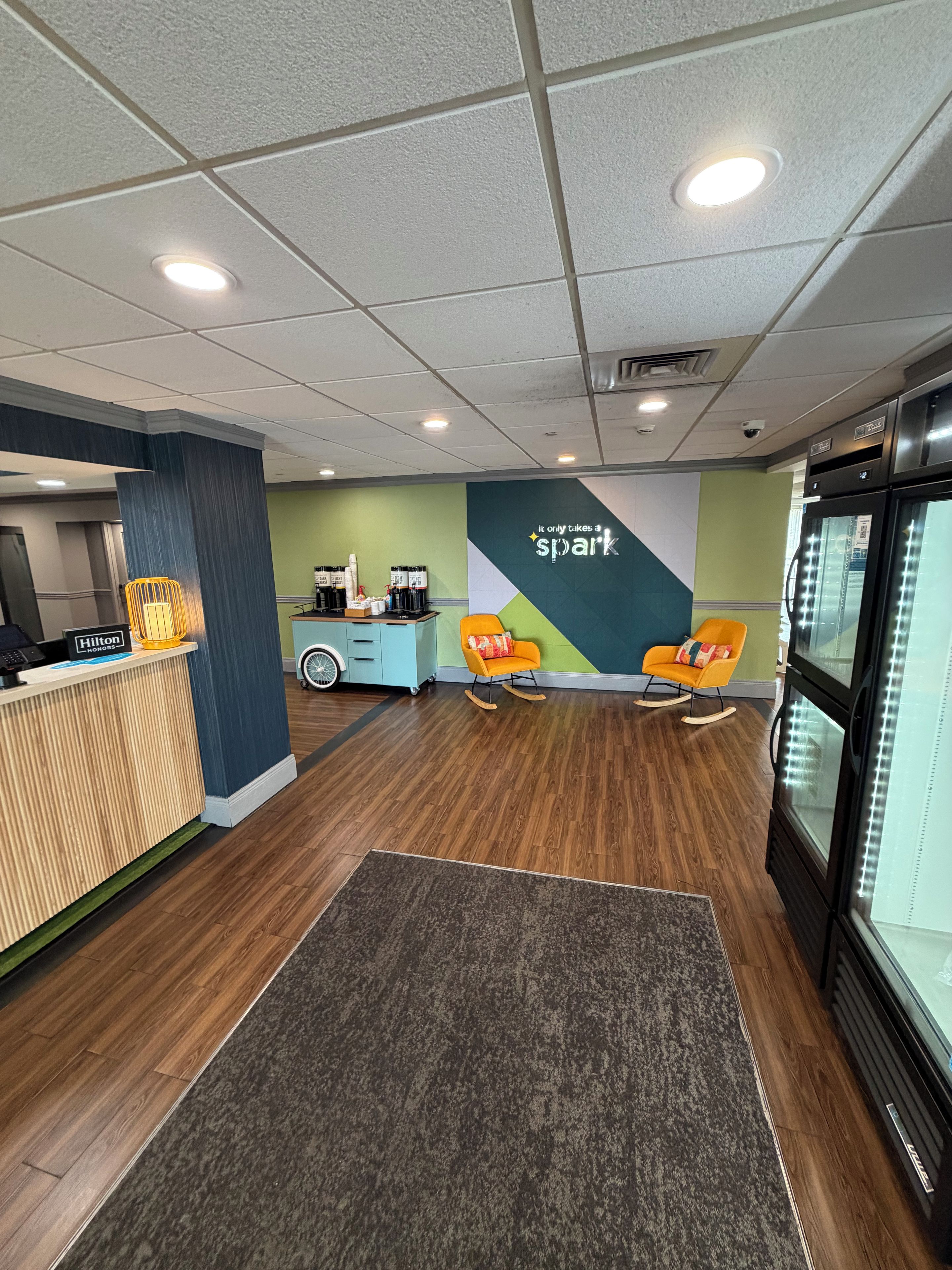 A hotel reception area with a wooden reception desk, a seating area with two rocking chairs, a wall with a motivational quote, and a refrigerator on the right.