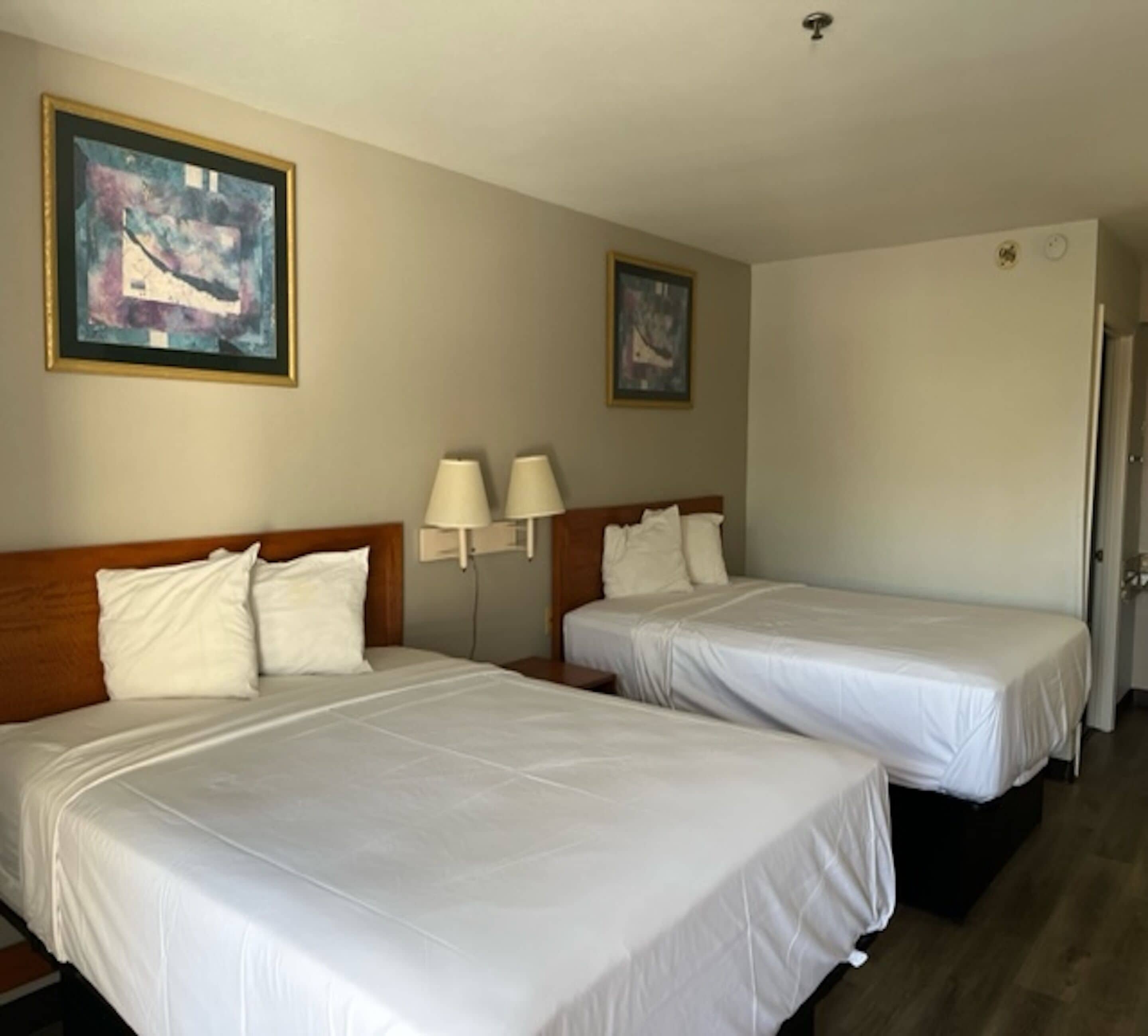 Habitació d'hotel amb dos llits, un panell de fusta al capçal, roba de llit blanca, dues làmpades de paret i dos quadres abstractes emmarcados.