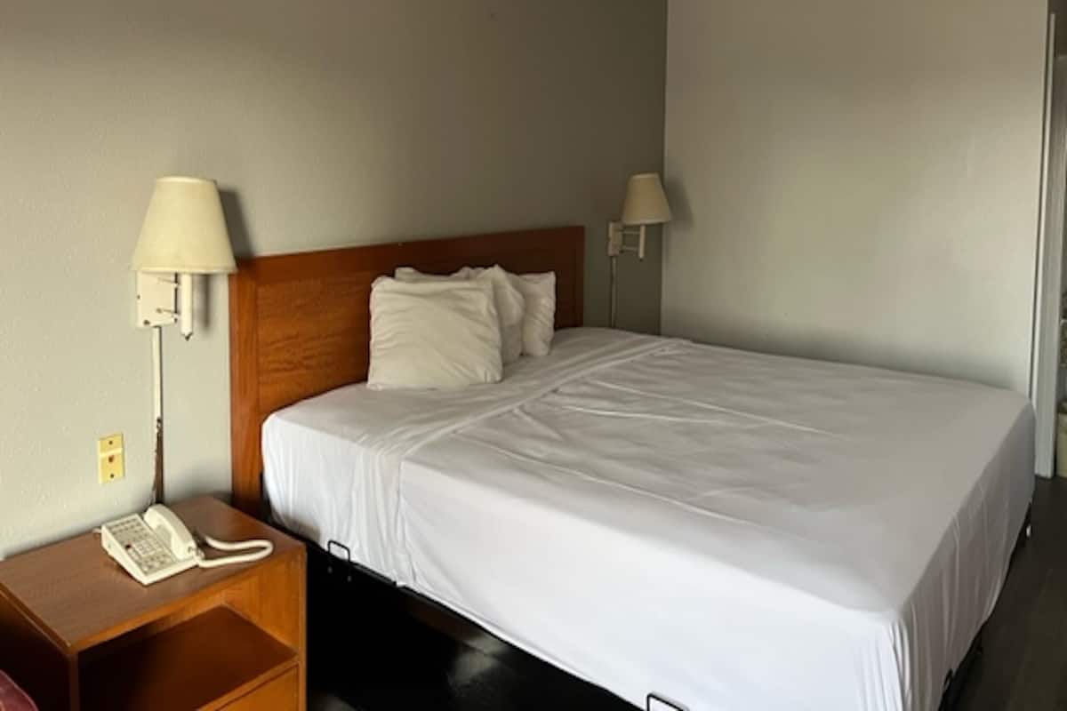 Habitación de hotel con una cama individual, cabecera de madera, dos lámparas de noche, una mesita de noche con teléfono y una pared lisa con interruptor de luz.