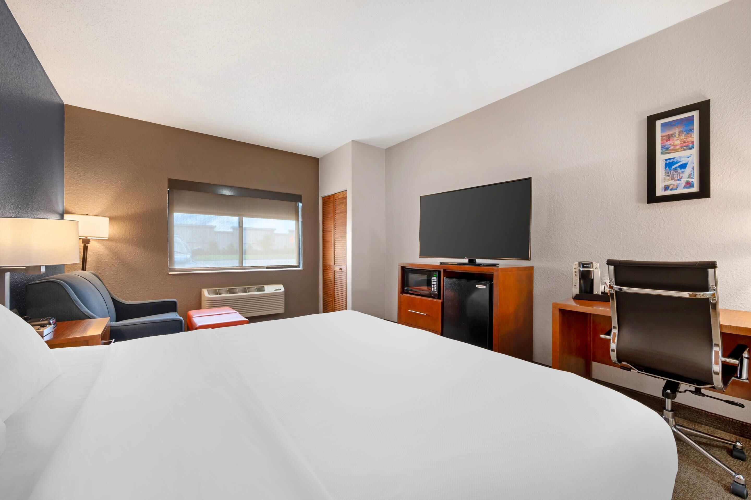 Quarto de hotel com uma cama grande, uma escrivaninha com computador, uma televisão e uma cadeira.