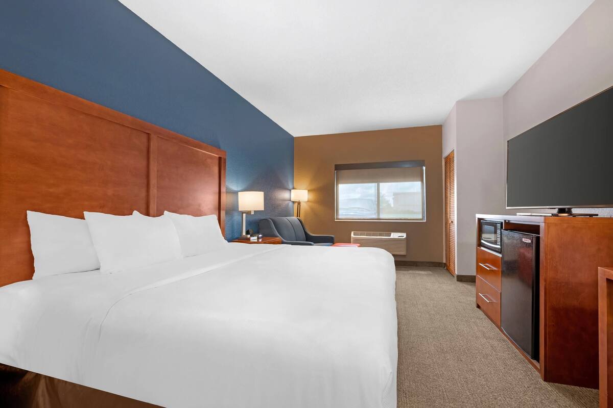 A hotel room with a large bed, a desk with a computer, a chair, a TV, and a window with blinds.