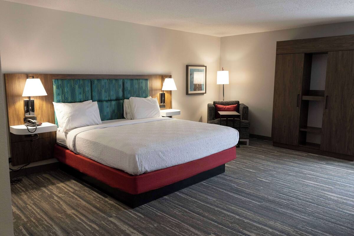 A hotel room with a large bed, two bedside tables with lamps, a wooden headboard, a red accent on the bed base, a small seating area with a chair and table, and a wardrobe.