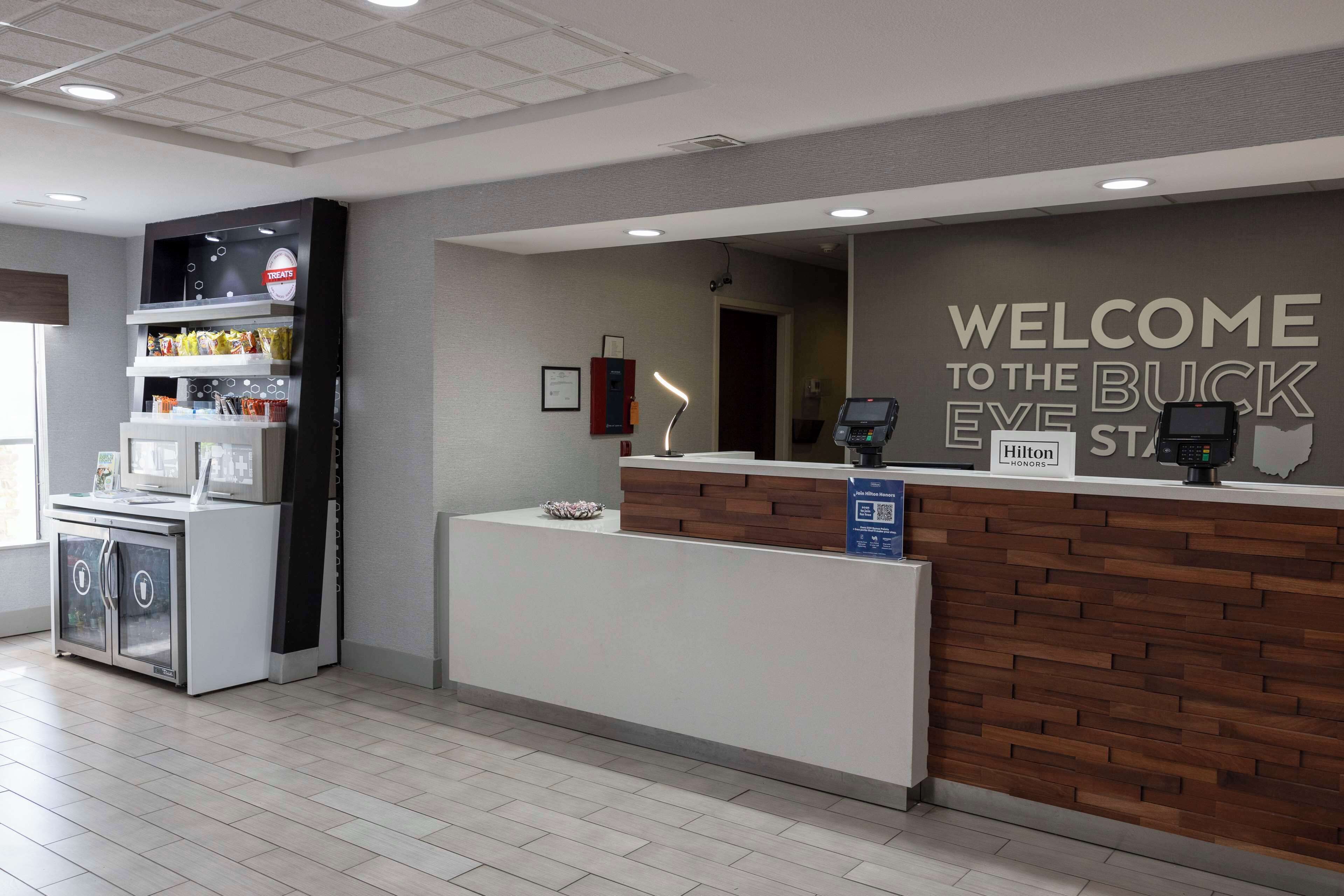 Zona de recepción de hotel con un diseño moderno, que incluye un mostrador de recepción, una pantalla digital y un cartel de bienvenida a los huéspedes en el Buck Eye State.