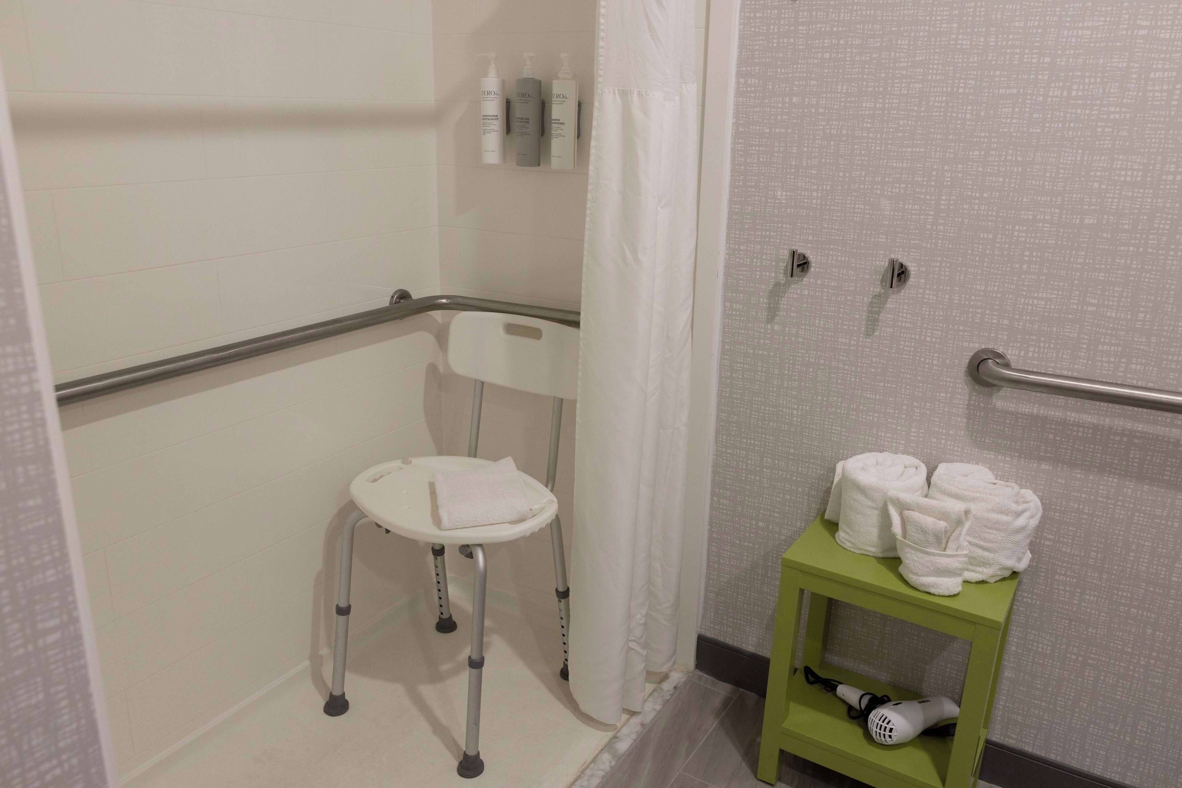 A bathroom with a wheelchair-accessible shower area, a small stool with a towel, and a shelf with toiletries and towels.